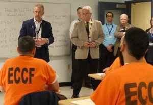 Two Essex County inmates wearing orange suits are in a classroom listening to NECC President Lane Glenn who is in front of the room with Essex County Sheriff Kevin Coppinger