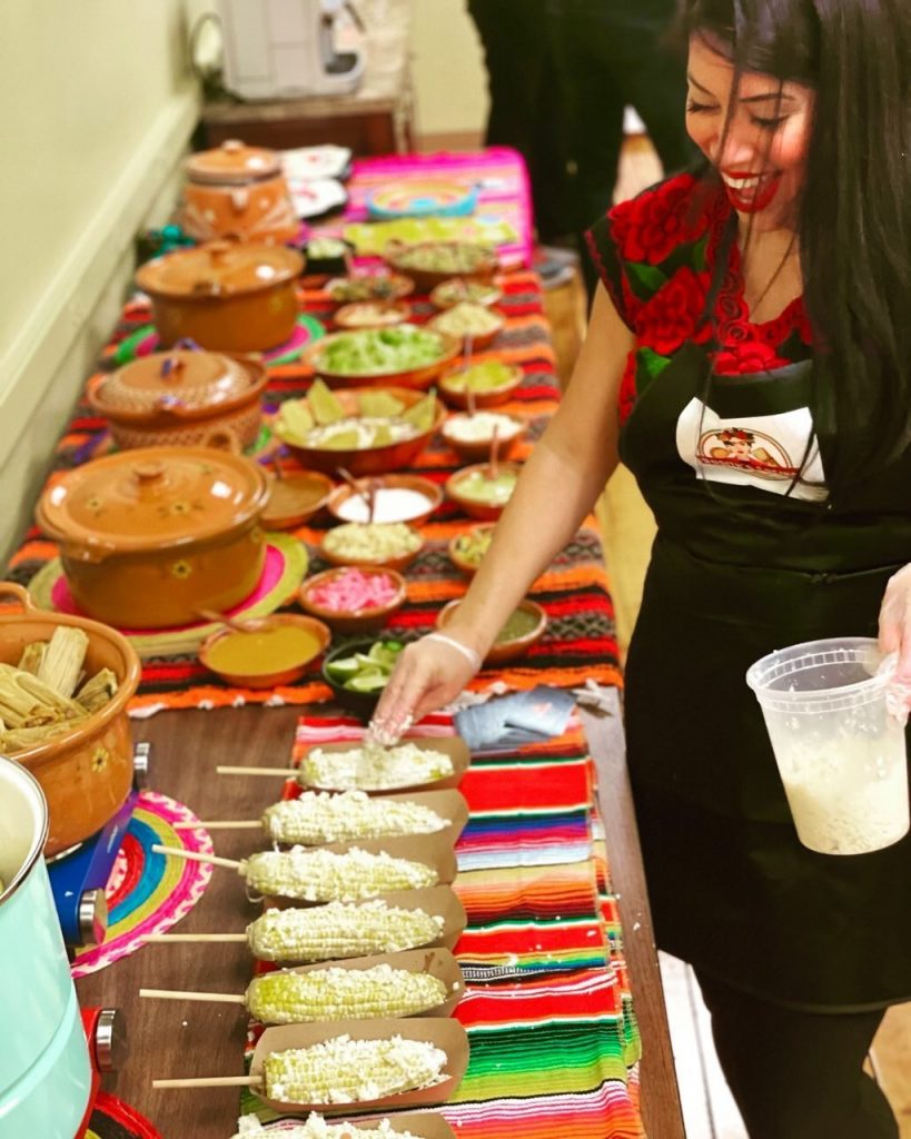 long table full of catered mexican dishes, rosa adds cheese to mexican street corn