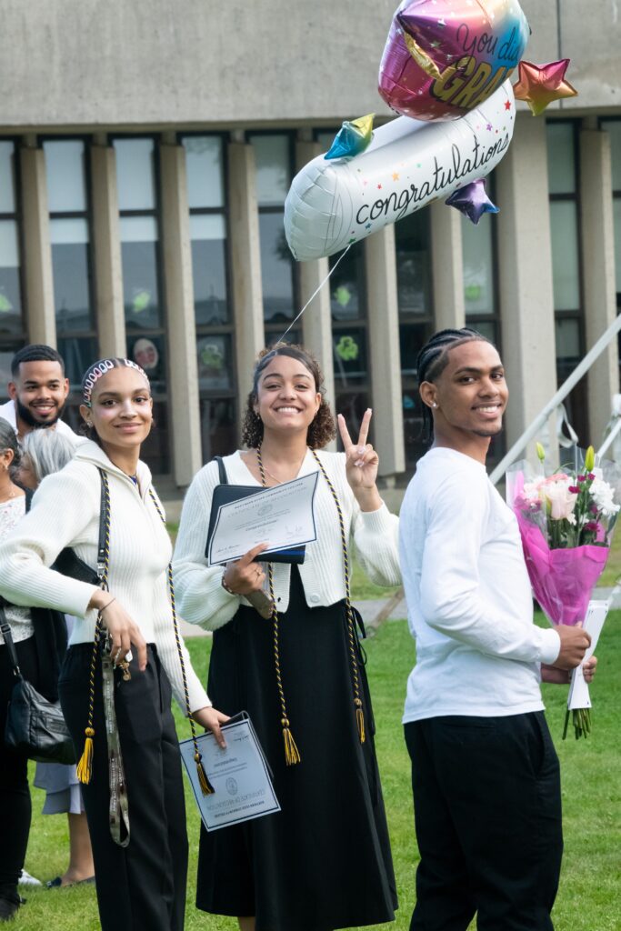 three students in white tops and black pants smile at the camera. one is holding a bunch of balloons with the word "congratulations" 