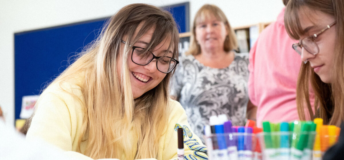 Two female presenting education students working on a group project together with markets while an instructor looks on from the background