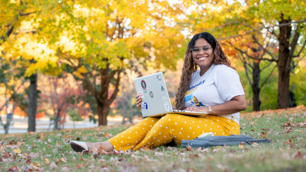 A photo of a student sitting on the NECC campus in the Fall with laptop in lap, smiling at the camera