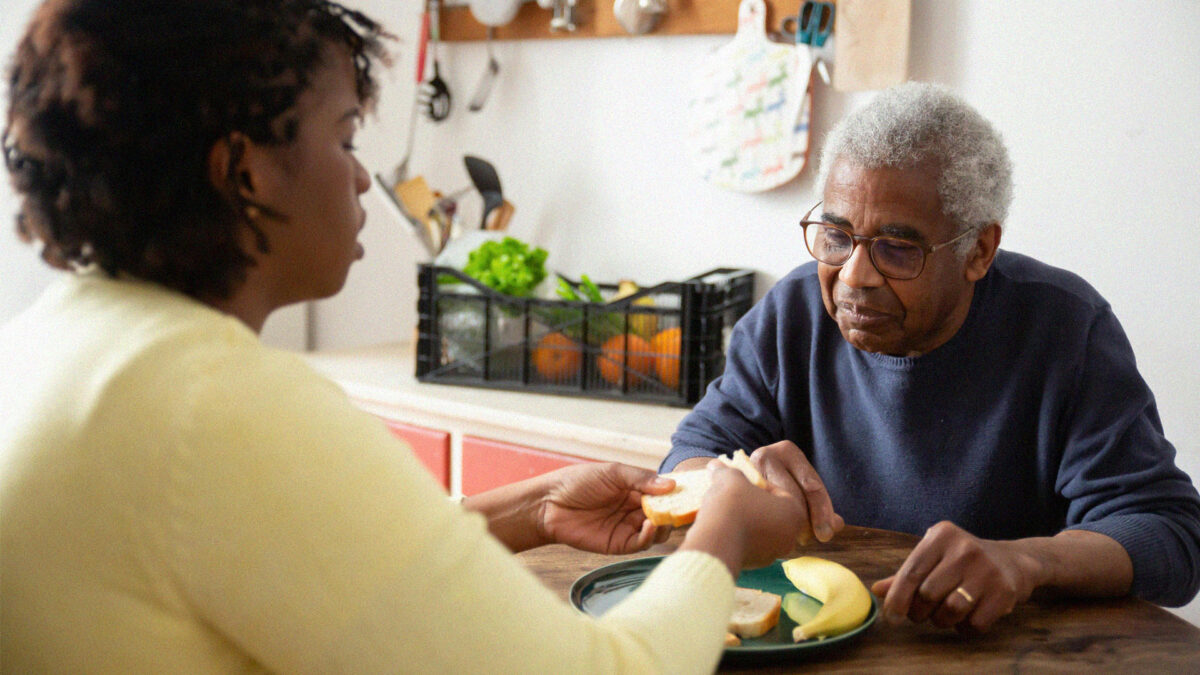 A young woman helping an older man prepare his food while sitting at a brightly lit table